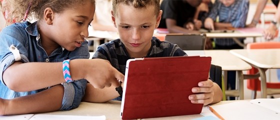 A boy and a girl in a classroom setting looking at a digital device / tablet.