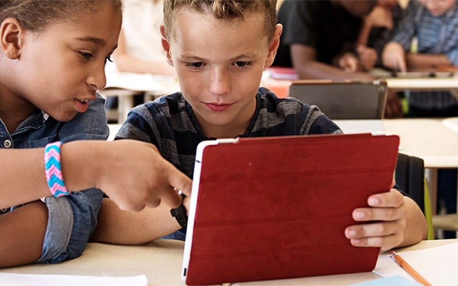 A boy and a girl in a classroom setting looking at a digital device / tablet.