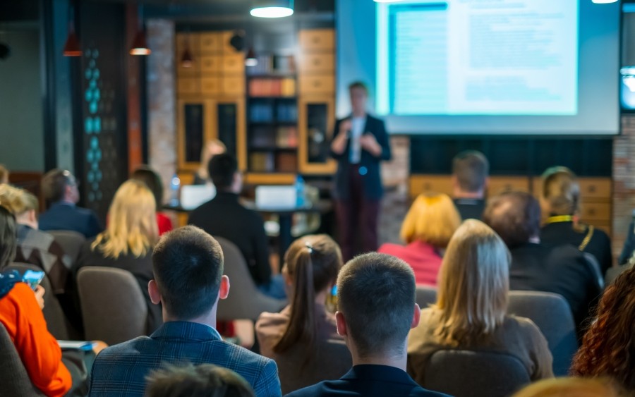 A seated crowd observes a speaker in front of a large digital screen with written information displayed.